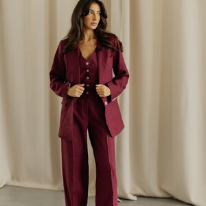 May include: A three-piece burgundy suit. The suit includes a blazer, a vest with button closures, and wide-leg trousers. The blazer has a classic lapel and side pockets. The outfit is made of a solid-colored fabric.