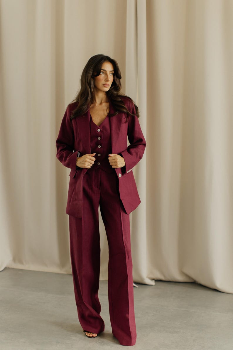 May include: A three-piece burgundy suit. The suit includes a blazer, a vest with button closures, and wide-leg trousers. The blazer has a classic lapel and side pockets. The outfit is made of a solid-colored fabric.