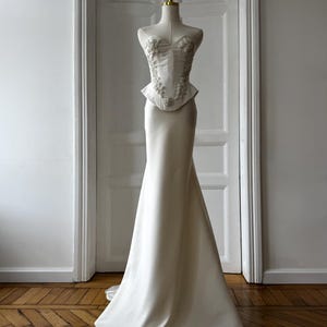 May include: A white wedding dress on a mannequin. The bodice features floral embellishments and a peplum waist, transitioning into a sleek, floor-length skirt. The dress is displayed in front of a white door and a wooden floor.