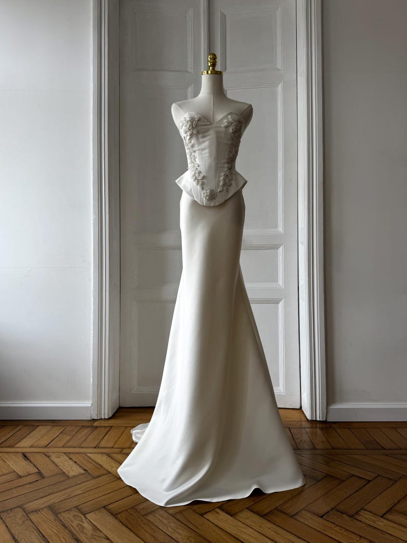 May include: A white wedding dress on a mannequin. The bodice features floral embellishments and a peplum waist, transitioning into a sleek, floor-length skirt. The dress is displayed in front of a white door and a wooden floor.