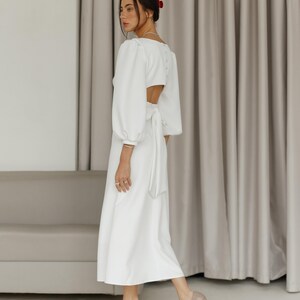May include: A white dress with a long sleeve, a cinched waist, and a low back. The dress is made of a lightweight fabric and has a flowing silhouette.