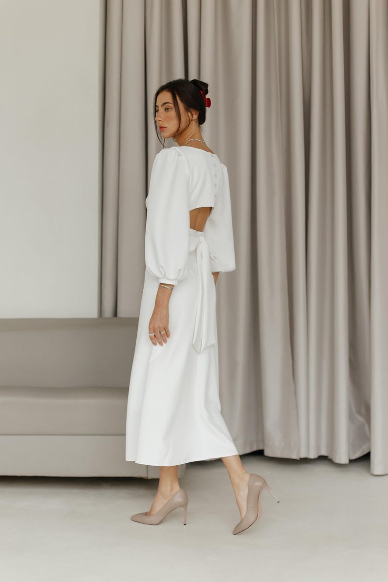 May include: A white dress with a long sleeve, a cinched waist, and a low back. The dress is made of a lightweight fabric and has a flowing silhouette.