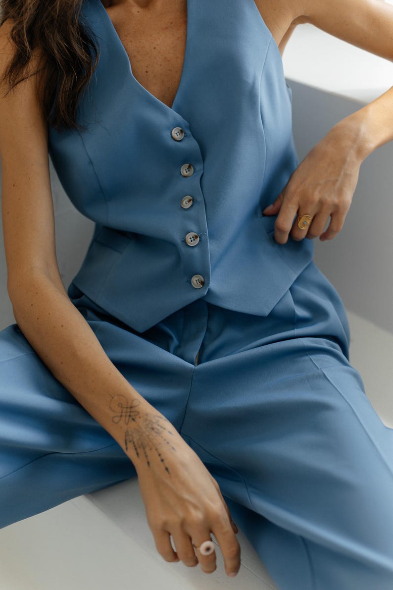 May include: A woman wearing a light blue pantsuit with a vest and matching pants. The vest has a V-neck and five buttons down the front. The woman is sitting with her legs crossed and her right hand resting on her left thigh.