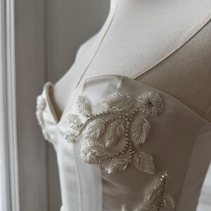 May include: Ivory-colored satin bodice with delicate floral beadwork and sparkling rhinestone accents. The bustier-style top features a sweetheart neckline and thin straps, creating an elegant and romantic look. The design is displayed on a mannequin.