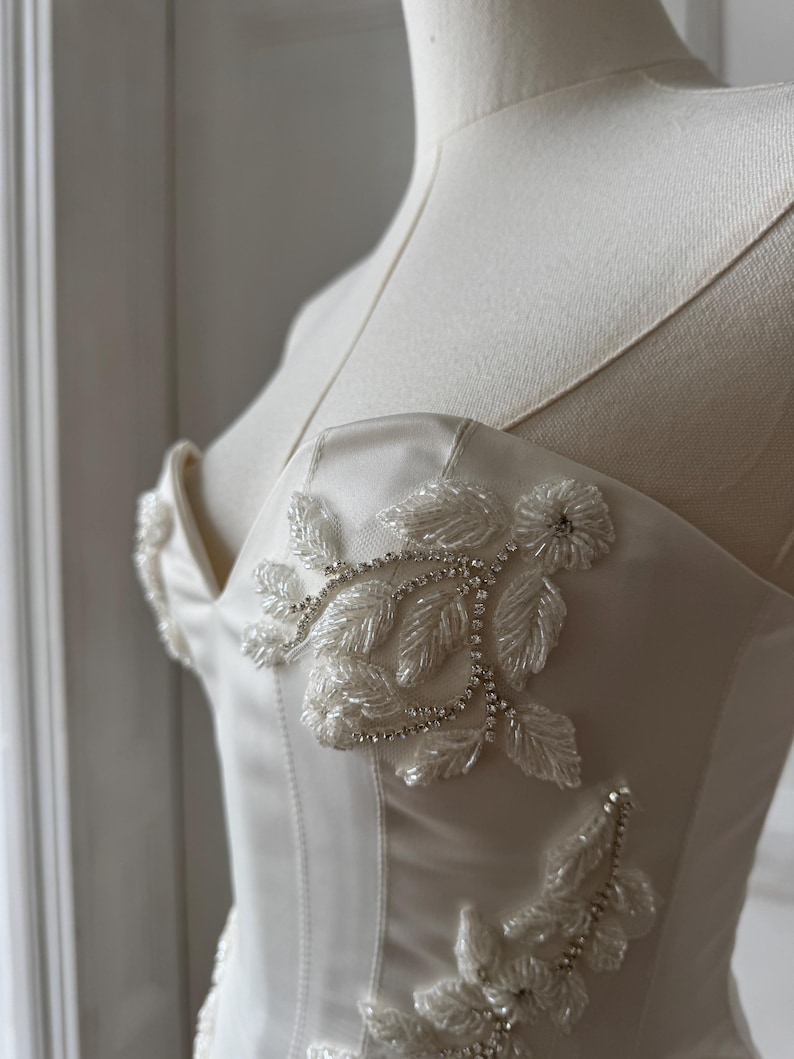 May include: Ivory-colored satin bodice with delicate floral beadwork and sparkling rhinestone accents. The bustier-style top features a sweetheart neckline and thin straps, creating an elegant and romantic look. The design is displayed on a mannequin.