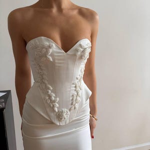May include: A strapless white mini dress with a structured bodice and a fitted skirt. The bodice features intricate floral embroidery with beaded accents. The dress is made of a smooth, satin-like fabric, creating a sleek and elegant look.