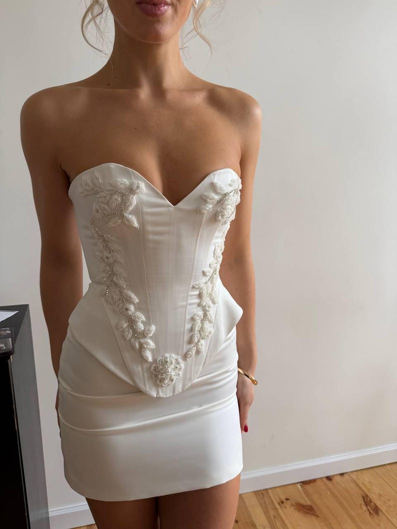 May include: A strapless white mini dress with a structured bodice and a fitted skirt. The bodice features intricate floral embroidery with beaded accents. The dress is made of a smooth, satin-like fabric, creating a sleek and elegant look.