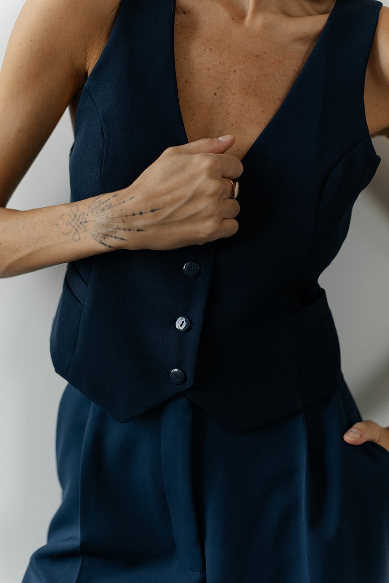 May include: A navy blue sleeveless vest with four buttons and a matching pair of pants. The vest has a deep V-neckline.