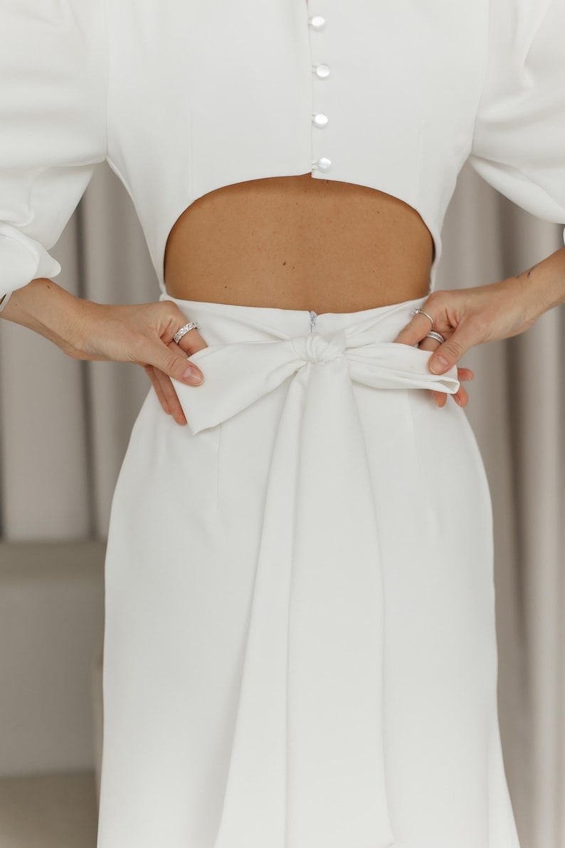 May include: White midi dress with a cutout at the waist and a bow tie at the back.