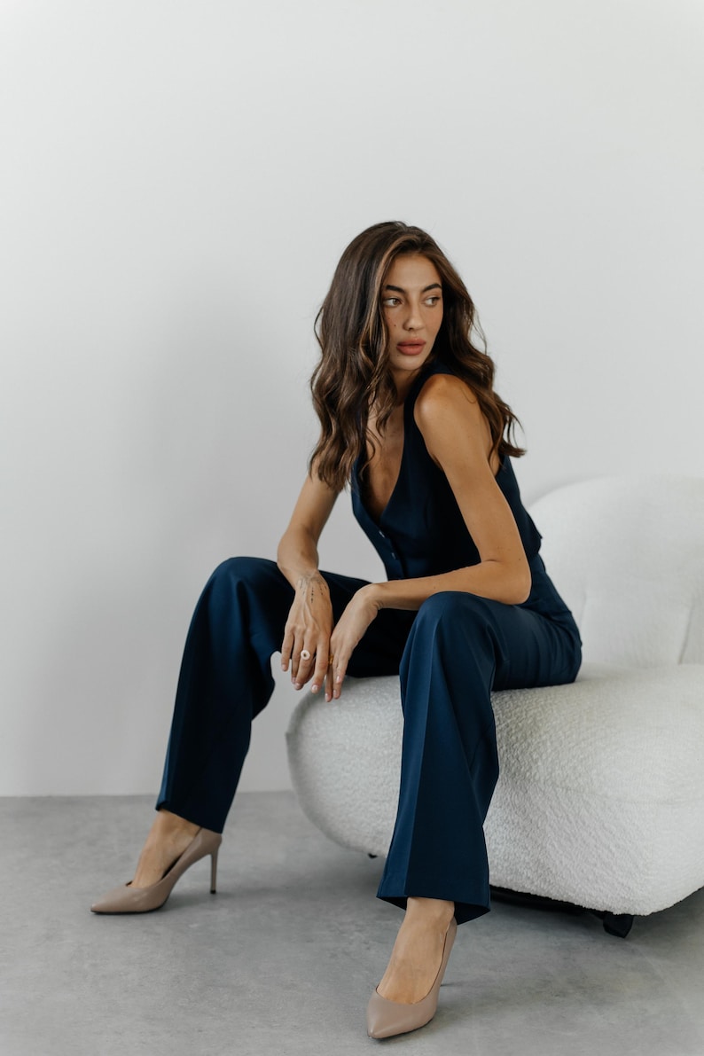 May include: A woman wearing a dark blue jumpsuit with a button-up front and wide-leg pants. She is sitting on a white fluffy chair and wearing nude heels.