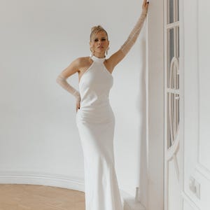 May include: A woman wearing a white halter neck dress with a fitted silhouette and a long skirt. She is standing in a white room with a white door behind her. She is wearing white gloves and white shoes.
