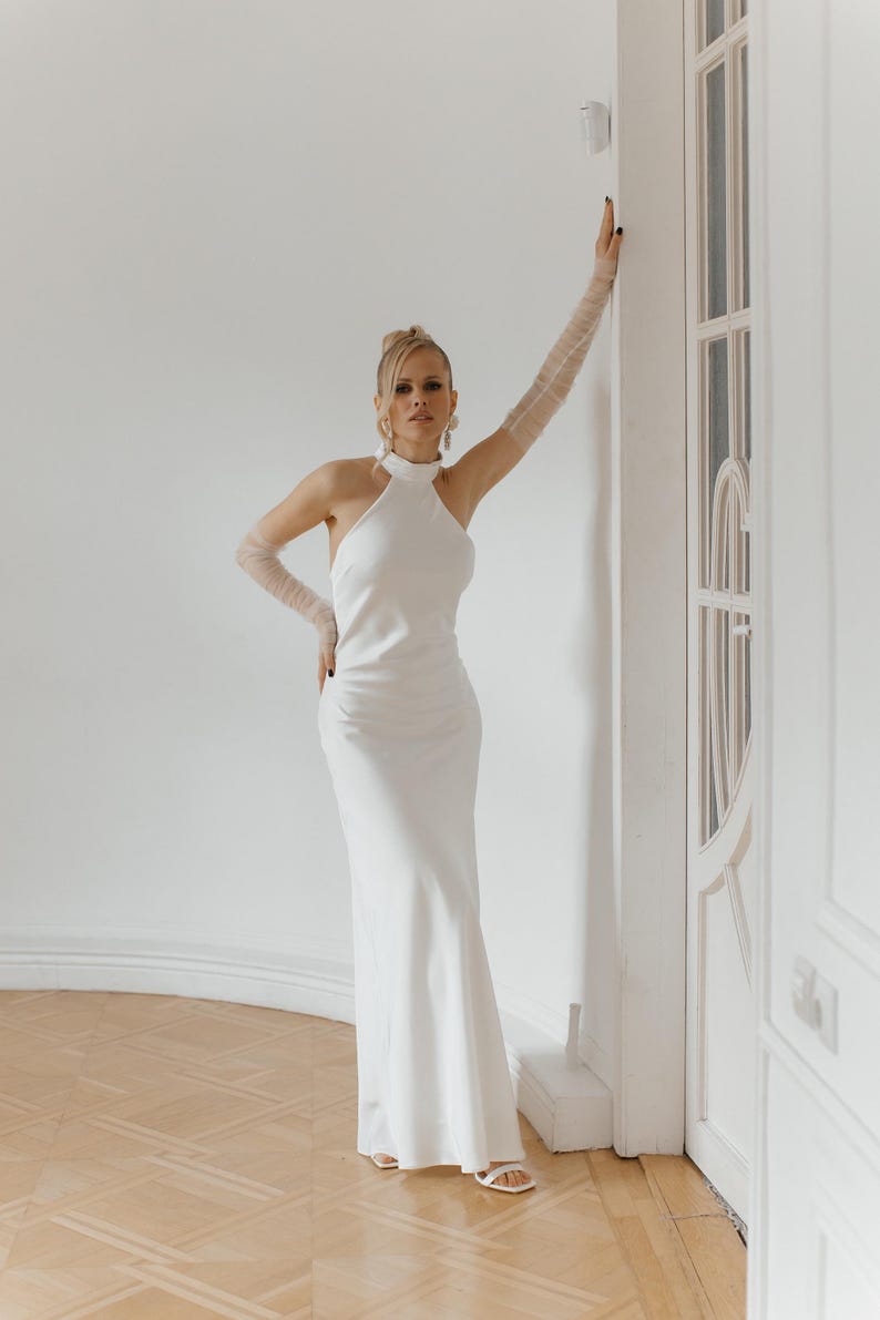 May include: A woman wearing a white halter neck dress with a fitted silhouette and a long skirt. She is standing in a white room with a white door behind her. She is wearing white gloves and white shoes.