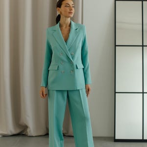 May include: A woman wearing a light blue double-breasted blazer and matching wide-leg pants. The blazer has four buttons and a pocket on each side. The pants have a straight leg.