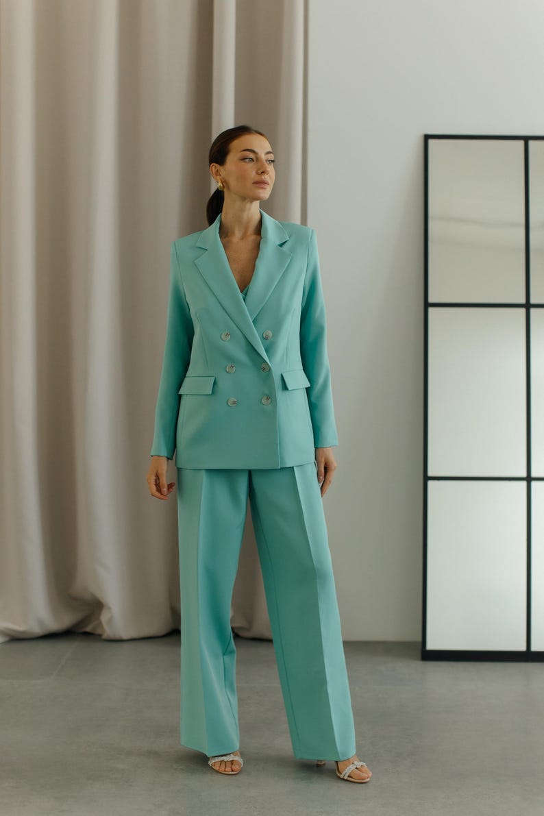 May include: A woman wearing a light blue double-breasted blazer and matching wide-leg pants. The blazer has four buttons and a pocket on each side. The pants have a straight leg.