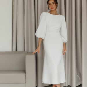 May include: A white midi dress with a fitted bodice and a flowing skirt. The dress has long sleeves with puffed shoulders and a round neckline.