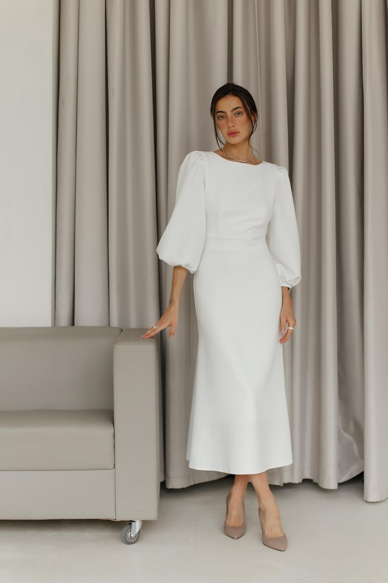 May include: A white midi dress with a fitted bodice and a flowing skirt. The dress has long sleeves with puffed shoulders and a round neckline.