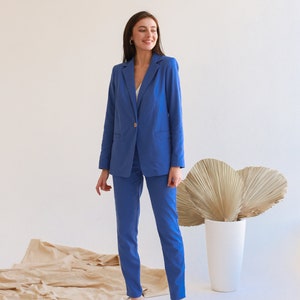 May include: A woman wearing a blue blazer and matching pants. She is standing in front of a white wall with a large, dried palm leaf behind her.