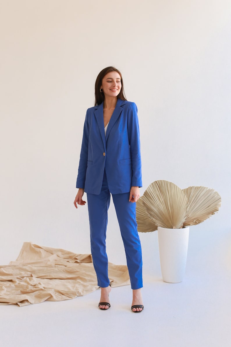 May include: A woman wearing a blue blazer and matching pants. She is standing in front of a white wall with a large, dried palm leaf behind her.