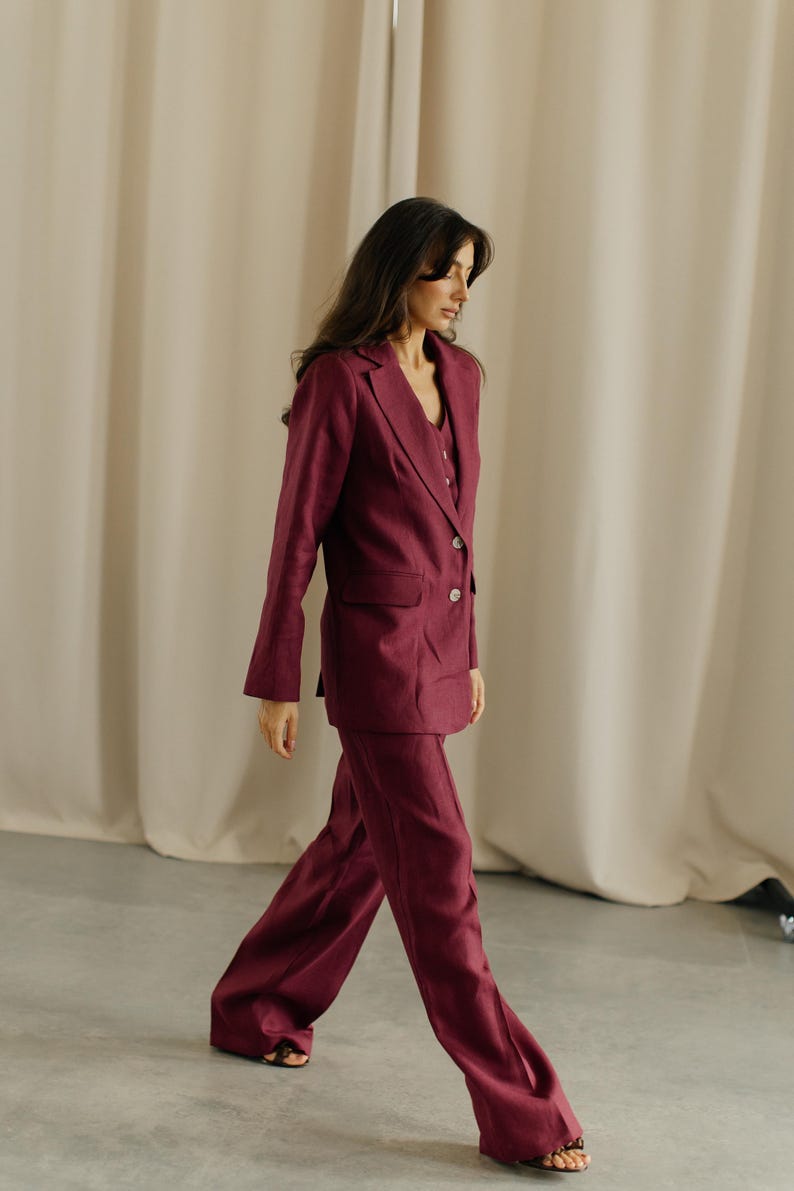May include: A burgundy linen suit, featuring a blazer with two buttons and wide-leg pants. The suit is a solid color and appears to be made of a lightweight fabric. The outfit is suitable for business or formal wear.