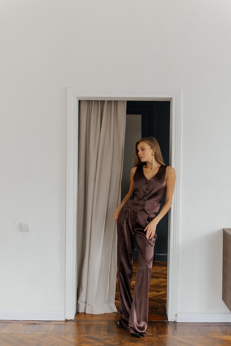 May include: A brown satin vest and matching wide-leg pants. The sleeveless vest has a V-neck and button closure. The pants have a high waist and a flowing silhouette. The outfit is a solid, rich brown color.