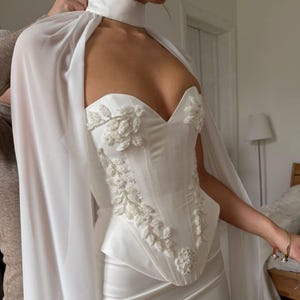 May include: A white bridal gown featuring a sweetheart neckline, embellished with floral beadwork. The gown includes a matching cape and a high collar. The dress is made of a satin-like material, creating a luxurious look.