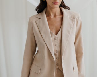 May include: A beige three-piece suit, including a blazer, vest, and trousers. The blazer has a notched lapel and two front pockets. The vest is buttoned up. The suit is made of a smooth fabric and is worn with gold hoop earrings.
