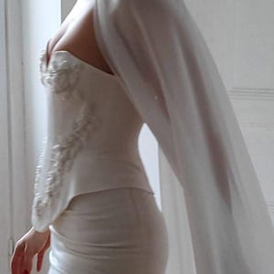 May include: A white, form-fitting dress with a sweetheart neckline and embellished bodice. The dress features a matching cape that drapes over the shoulder. The dress is knee-length and appears to be made of a smooth fabric.