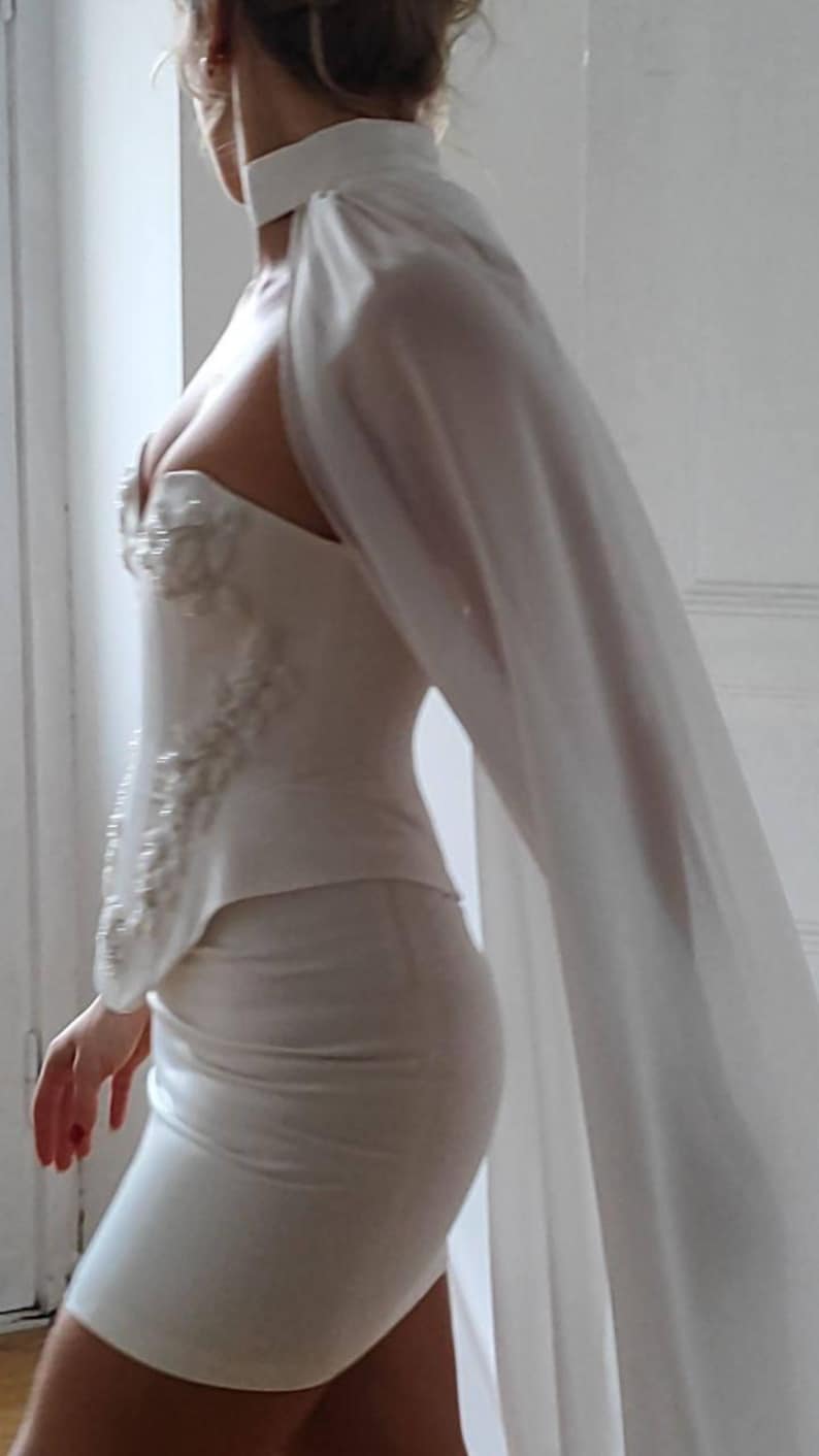 May include: A white, form-fitting dress with a sweetheart neckline and embellished bodice. The dress features a matching cape that drapes over the shoulder. The dress is knee-length and appears to be made of a smooth fabric.