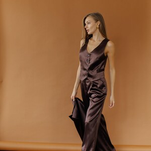 May include: A brown satin vest and matching wide-leg pants. The vest has a V-neck and button closure. The pants are high-waisted. The outfit is made of a shiny, flowing fabric. The model is wearing black shoes.