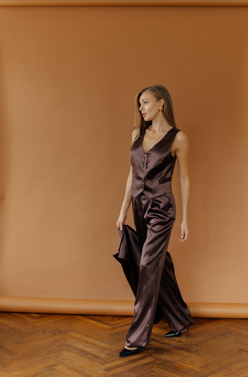 May include: A brown satin vest and matching wide-leg pants. The vest has a V-neck and button closure. The pants are high-waisted. The outfit is made of a shiny, flowing fabric. The model is wearing black shoes.