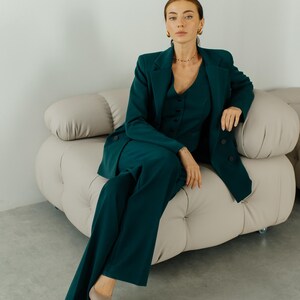 May include: A woman wearing a dark green pantsuit sits on a light beige sofa. She is wearing gold earrings and a gold necklace.