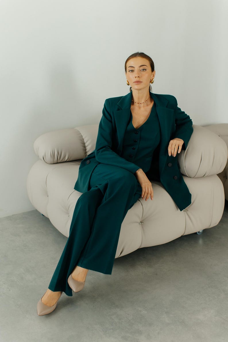 May include: A woman wearing a dark green pantsuit sits on a light beige sofa. She is wearing gold earrings and a gold necklace.