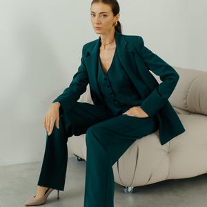 May include: A woman wearing a dark green pantsuit with a blazer and matching pants. She is sitting on a light beige couch with wheels. She is wearing beige heels.