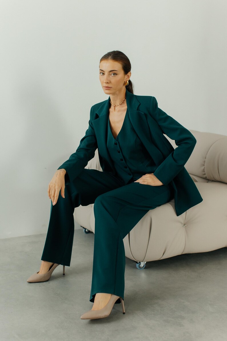 May include: A woman wearing a dark green pantsuit with a blazer and matching pants. She is sitting on a light beige couch with wheels. She is wearing beige heels.