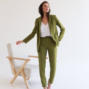 May include: A woman wearing a green pantsuit with a white top and black heels. She is standing in front of a white chair with wooden legs.