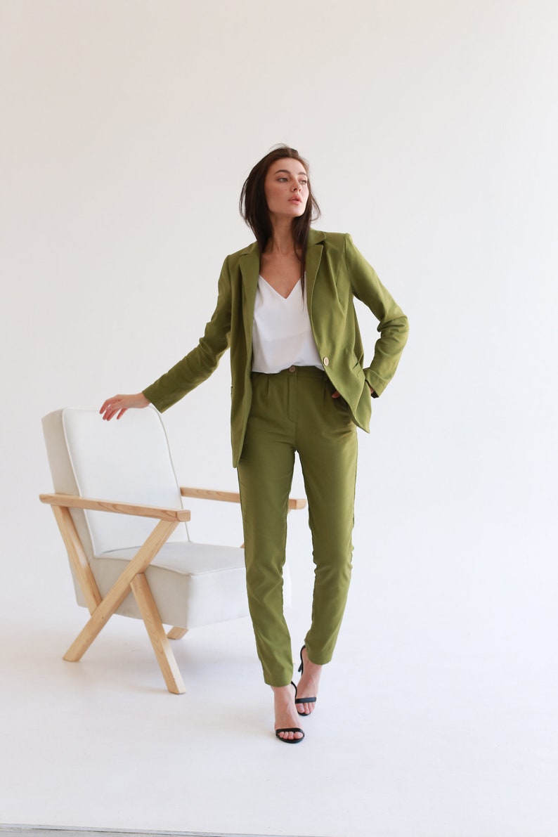 May include: A woman wearing a green pantsuit with a white top and black heels. She is standing in front of a white chair with wooden legs.