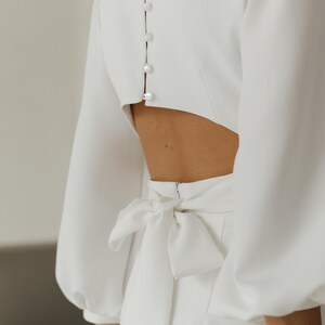 May include: A white dress with a low back and a bow tie at the waist. The dress has long sleeves with puffed shoulders and a button closure down the back.