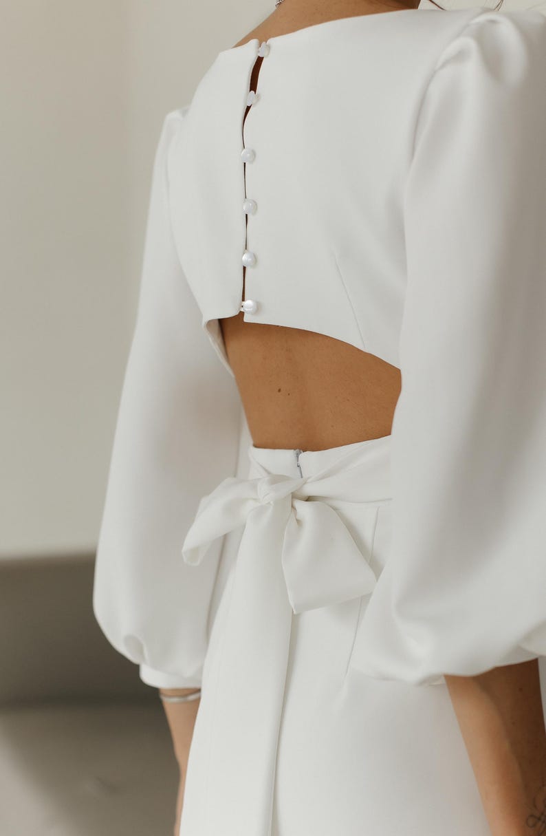 May include: A white dress with a low back and a bow tie at the waist. The dress has long sleeves with puffed shoulders and a button closure down the back.