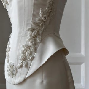 May include: Ivory-colored bridal gown with floral beaded embellishments. The bodice features a structured design with a fitted waist and a flared peplum detail. The intricate beadwork forms delicate floral patterns along the bodice.