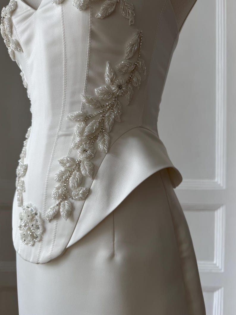 May include: Ivory-colored bridal gown with floral beaded embellishments. The bodice features a structured design with a fitted waist and a flared peplum detail. The intricate beadwork forms delicate floral patterns along the bodice.