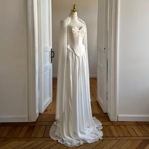 May include: A white bridal gown on a mannequin. The dress has a fitted bodice with floral detailing and a flowing skirt. A sheer cape is draped over the shoulders. The dress is displayed in a room with wooden flooring and white doors.