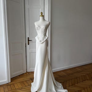 May include: A white wedding dress on a mannequin. The dress features a fitted bodice with floral embellishments and a flowing skirt that flares out at the bottom. The dress is displayed in a room with wooden floors and white doors.