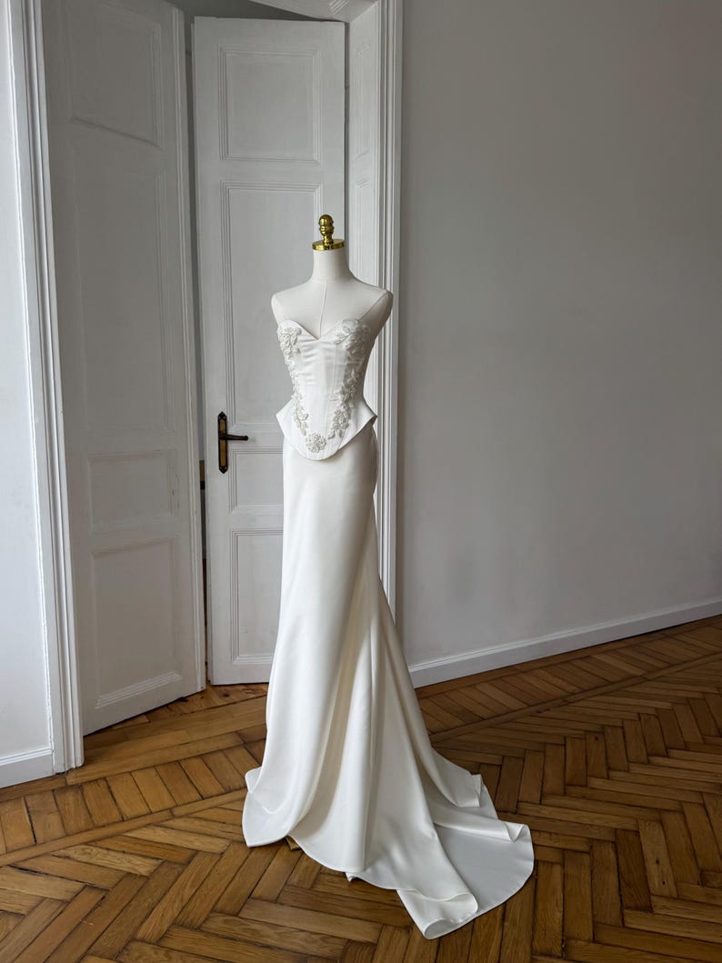 May include: A white wedding dress on a mannequin. The dress features a fitted bodice with floral embellishments and a flowing skirt that flares out at the bottom. The dress is displayed in a room with wooden floors and white doors.