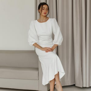 May include: A woman wearing a white dress with a fitted bodice and a flowing skirt. She is sitting on a white couch with her legs crossed and her hands in her lap. She is wearing nude heels.