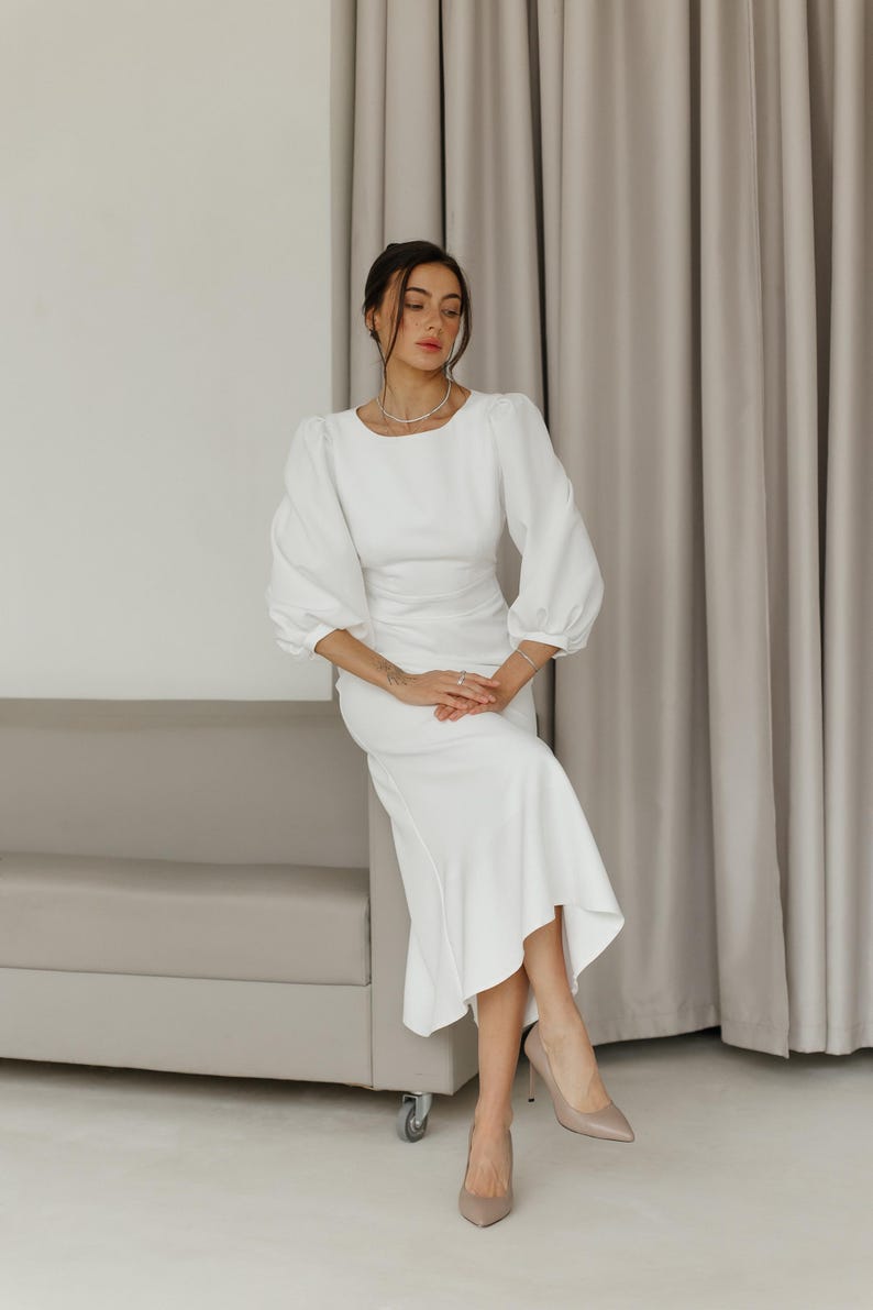 May include: A woman wearing a white dress with a fitted bodice and a flowing skirt. She is sitting on a white couch with her legs crossed and her hands in her lap. She is wearing nude heels.