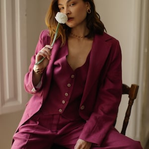 May include: A woman wearing a magenta suit with a vest and a single white flower tucked behind her ear. She is sitting on a wooden chair with a black seat.