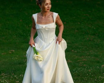 May include: A white satin wedding dress with a square neckline and a bow detail on the bodice. The dress has a full skirt and is being held up, revealing the wearer's feet. The person is standing in a grassy field and holding a bouquet of white flowers.