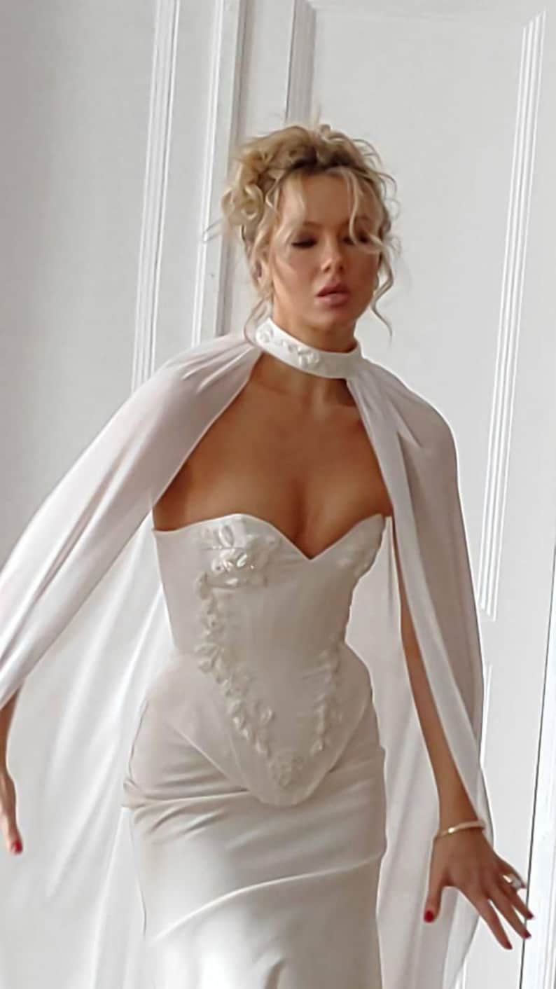 May include: A white bridal gown with a sweetheart neckline and floral embellishments. The dress features a fitted bodice and a flowing skirt. A matching cape is draped over the shoulders, and a choker necklace is worn around the neck.