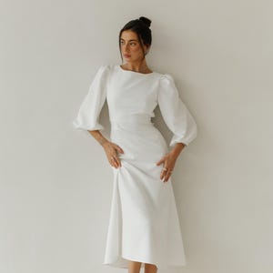 May include: A woman wearing a white dress with a fitted bodice and a flowing skirt. The dress has long sleeves with puffed shoulders. She is wearing nude heels.