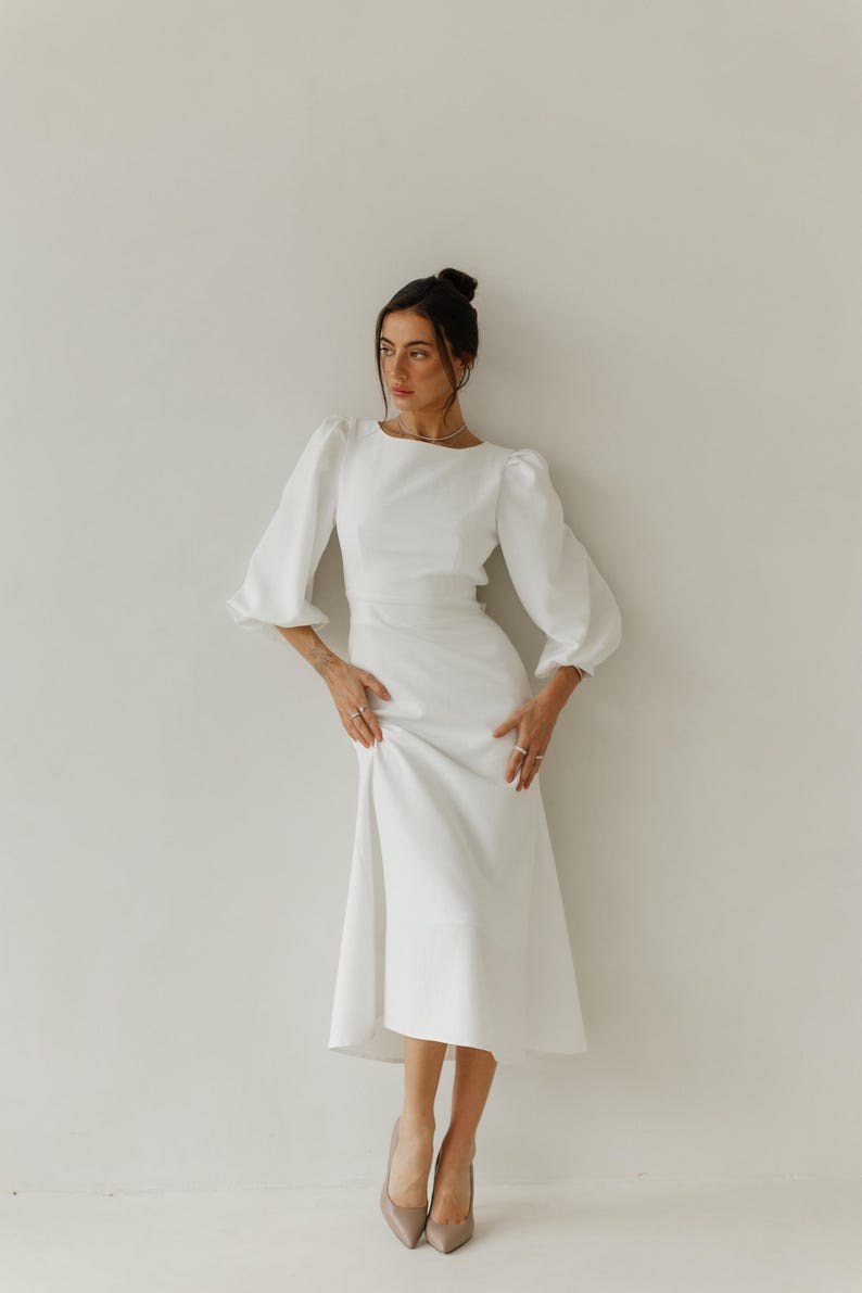 May include: A woman wearing a white dress with a fitted bodice and a flowing skirt. The dress has long sleeves with puffed shoulders. She is wearing nude heels.